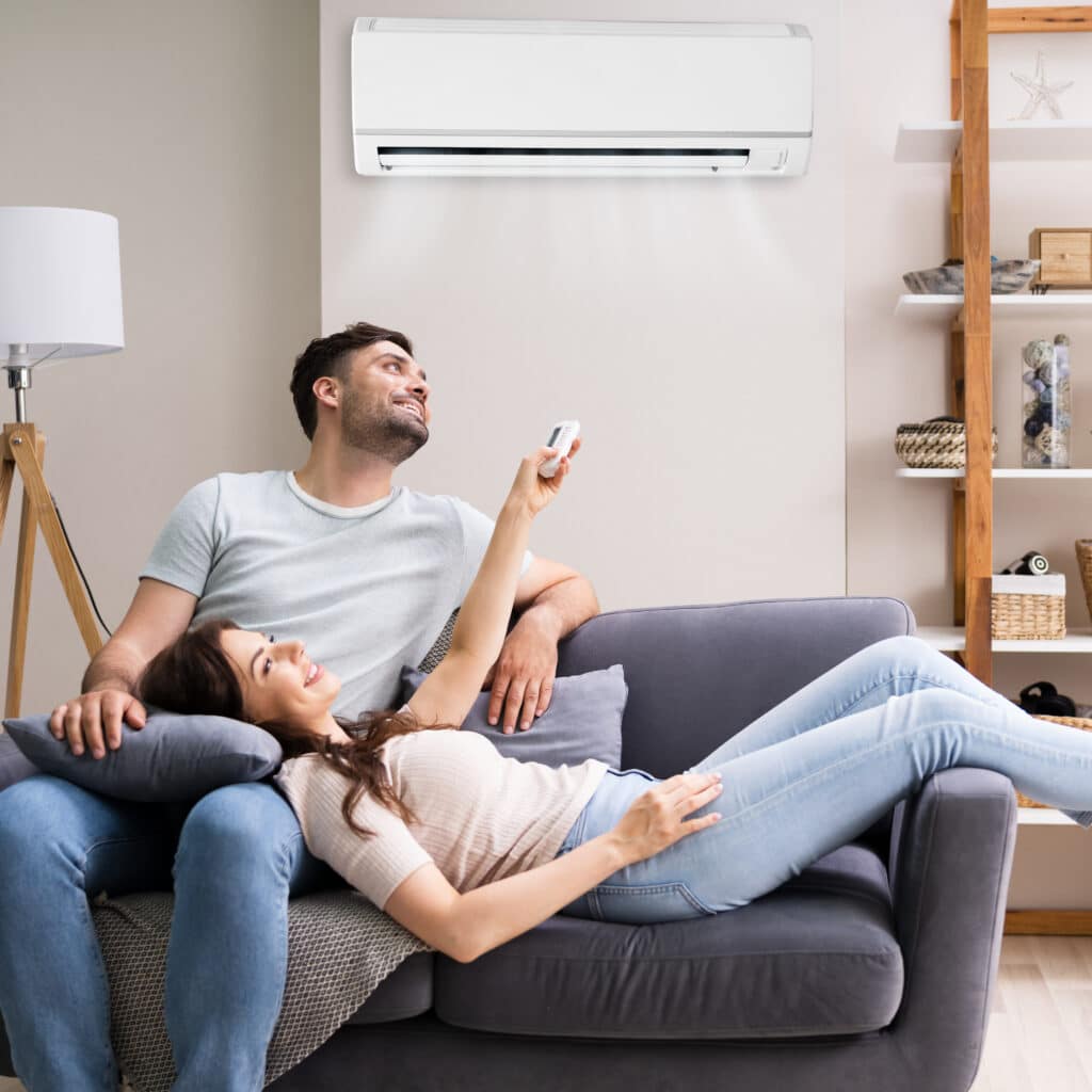 Couple : confort et fraîcheur à la maison avec climatiseur Couple souriant sur un canapé, la femme allongée sur l'homme qui règle un climatiseur mural avec une télécommande.