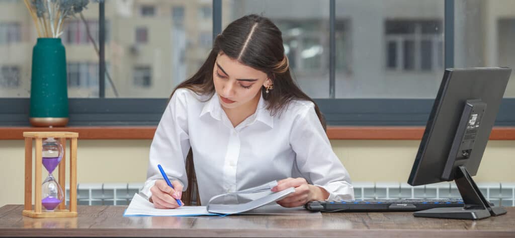Femme au bureau: Travail, Étude et Concentration Jeune femme concentrée, en chemise blanche, écrit sur son bureau près d'un sablier violet et d'un écran d'ordinateur.