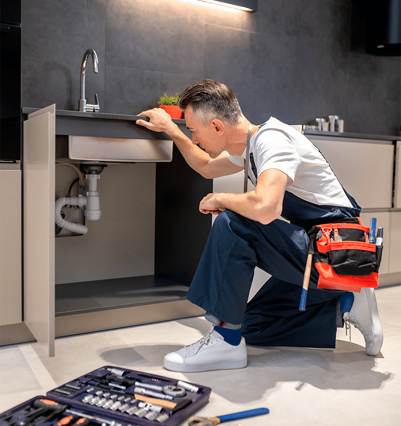 Plombier inspectant les tuyaux sous un évier de cuisine moderne. Il est accroupi, ses outils de dépannage sont au sol.