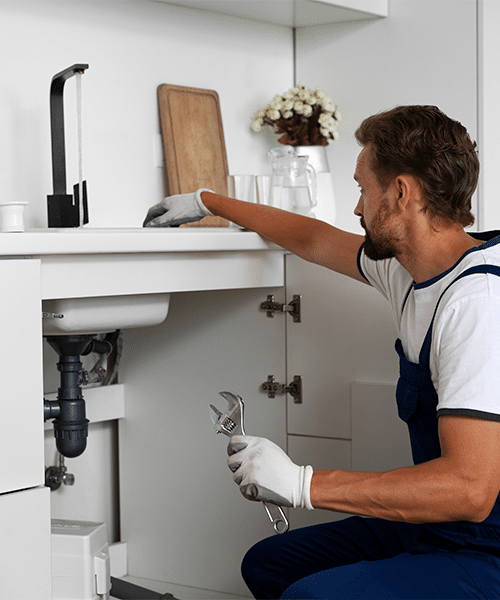 Plombier expert: Réparation d'évier et robinetterie de cuisine Plombier ganté en salopette bleue réparant les tuyaux sous un évier de cuisine moderne avec une clé. Robinet noir visible.