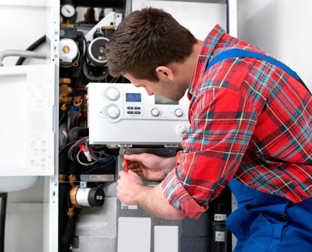 Réparation chaudière gaz : entretien professionnel et dépannage Technicien en chemise à carreaux rouge réparant une chaudière à gaz blanche. Il ajuste un composant avec un tournevis, affichage numérique 53 visible. Entretien de chauffage.